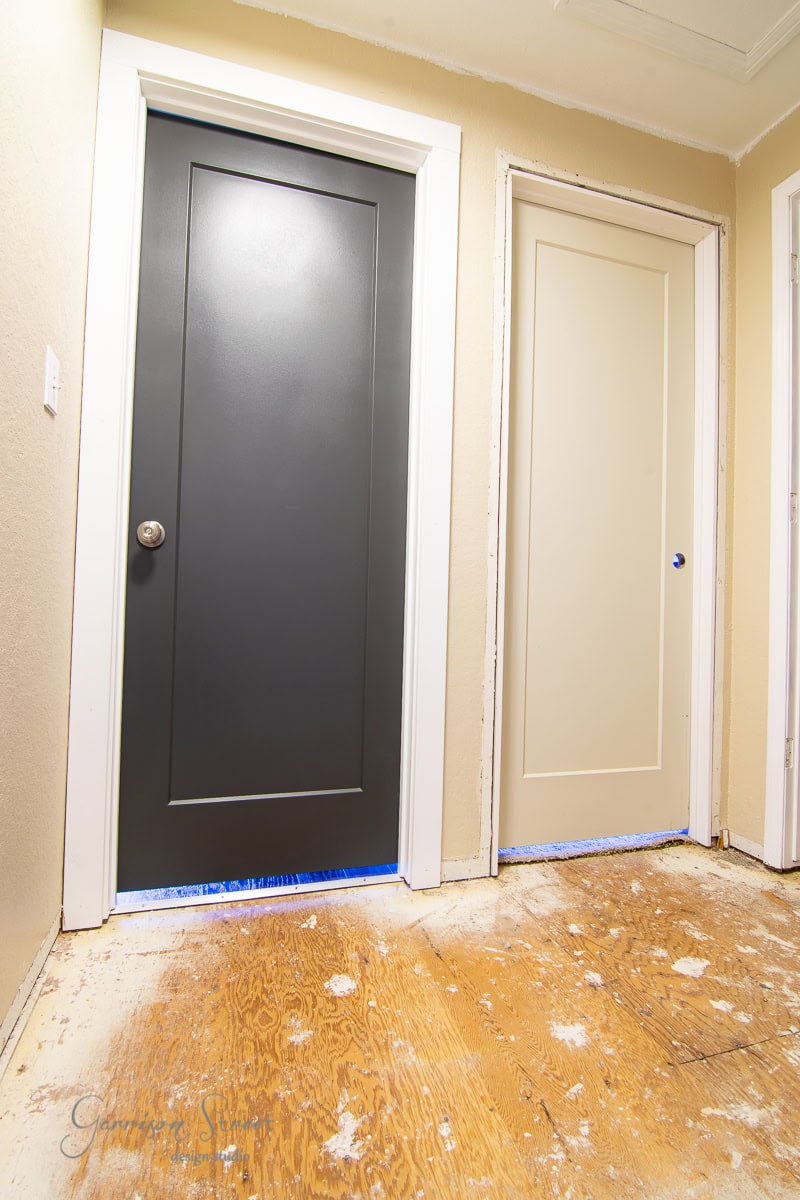 A hallway features two doors, one painted black and the other beige, both with white trim. The floor remains unfinished with visible plywood.