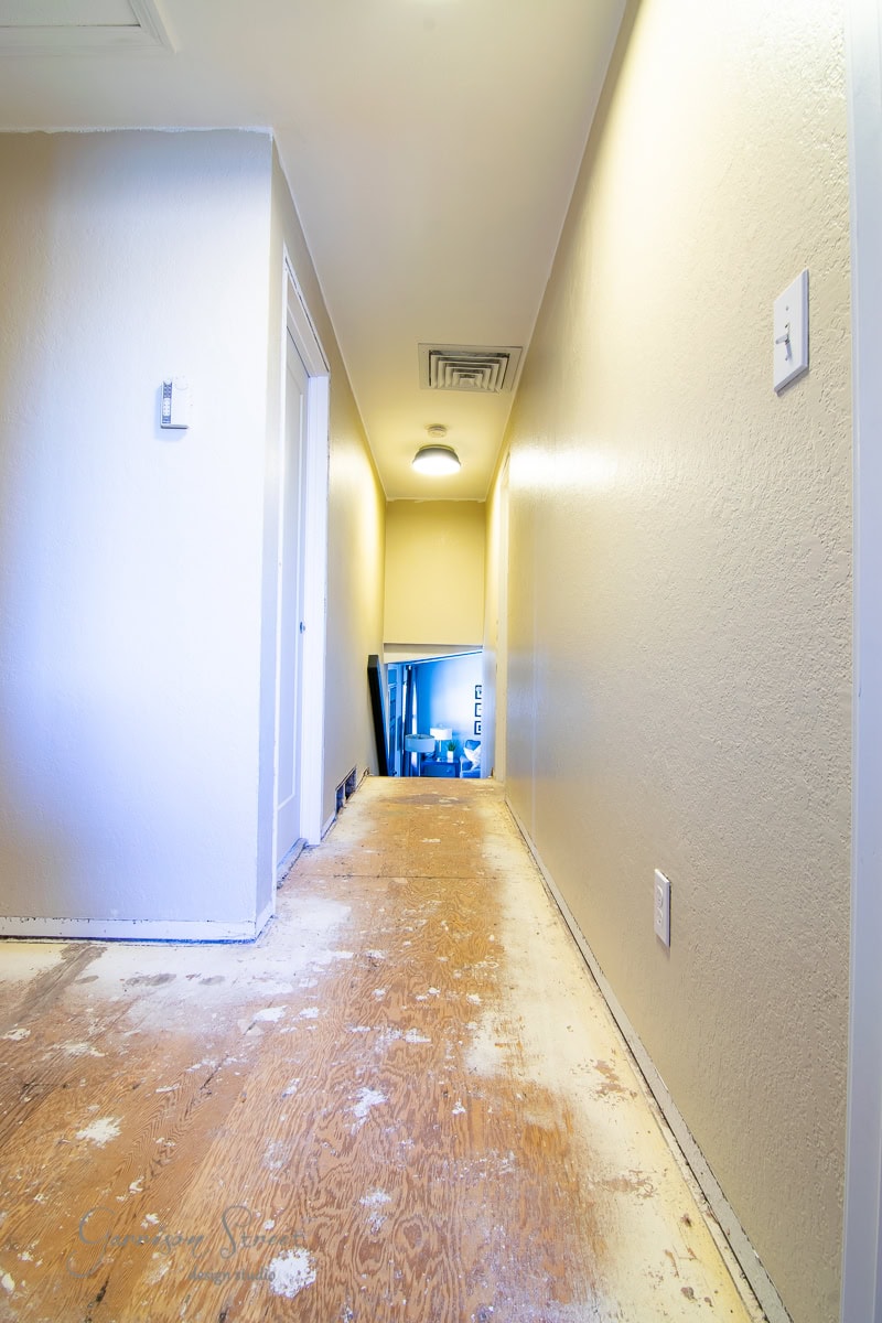 A narrow hallway with off-white walls and a bare wooden floor is under renovation. At the end, there's a doorway leading to a staircase with a handrail. Ceiling lights illuminate the space.