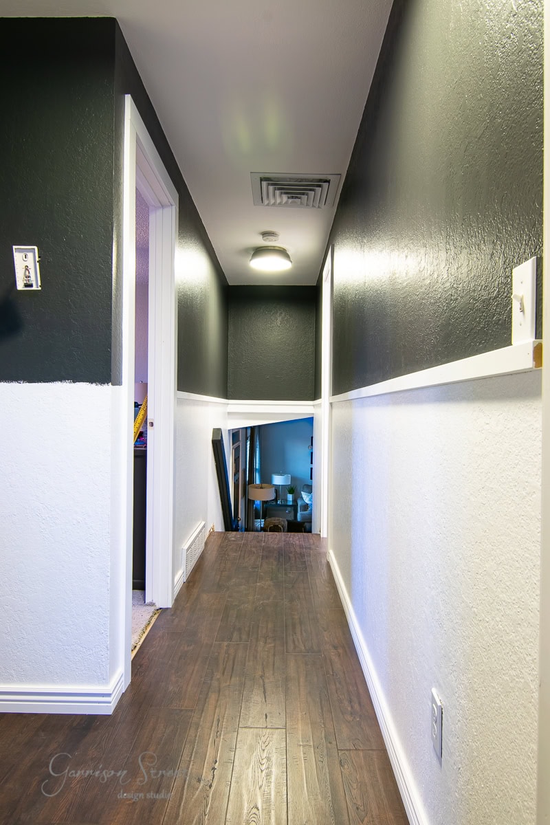 A narrow hallway features a board and batten wall design with dark upper walls and white lower sections, illuminated by ceiling lights. The dark wood floor complements the aesthetic, with visible doorways on the sides. At the end of the hallway, a lit room is partially visible.