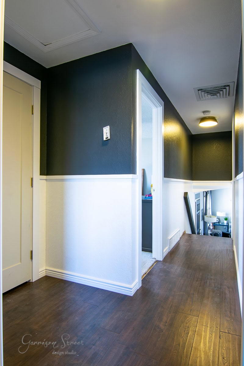 A hallway with DIY board and batten walls showcases white lower panels contrasting against dark upper walls, all complemented by a wooden floor. A round ceiling light brightens the space, while a door on the left and an open doorway at the end lead to another room.
