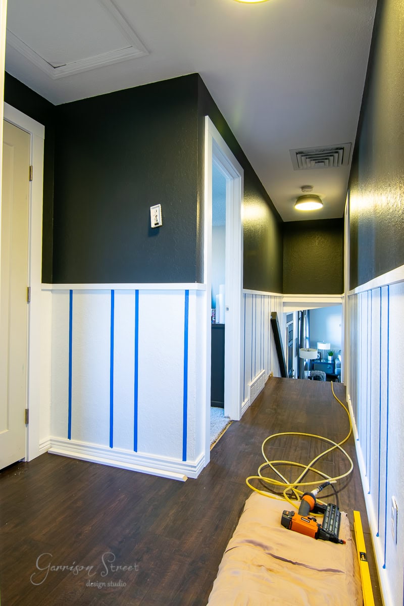 A hallway under renovation features dark walls and white board and batten wainscoting. Blue painter's tape lines the lower walls. A wooden floor contrasts with a yellow power cord and tools on a cloth. A partially open door is visible at the end.