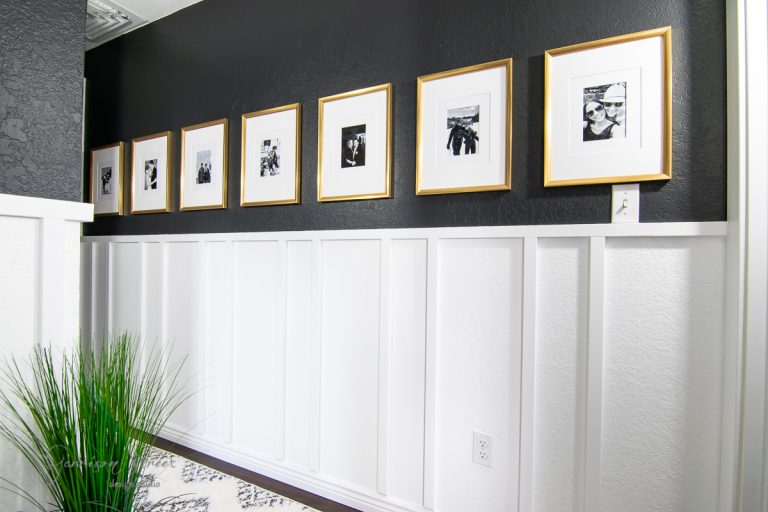 A hallway with dark walls and white wainscoting, reminiscent of a board and batten wall DIY project, is adorned with framed black-and-white photos in gold frames. Green grass-like plants add a pop of color, while a patterned rug covers the floor.