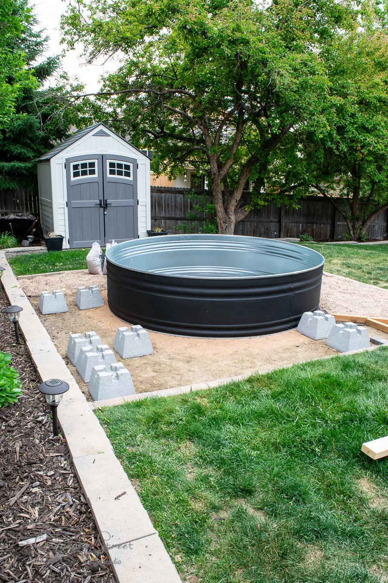 A backyard features a large, round, black stock tank pool set on a sand base, surrounded by concrete blocks. A gray shed with white trim stands nearby, framed by green trees and grass.