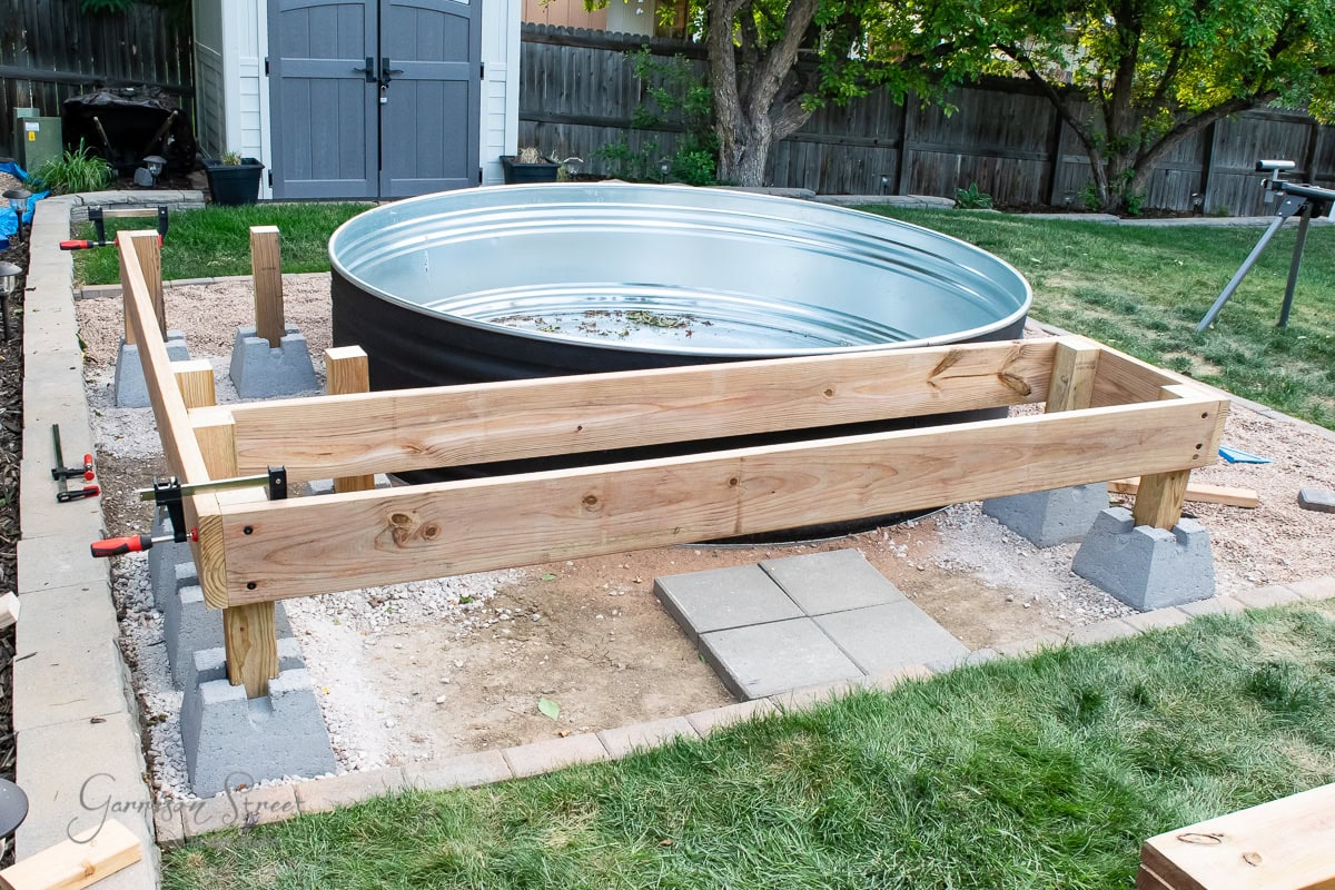 A metal stock tank surrounded by a partially constructed wooden frame in a backyard. The frame is supported by cinder blocks and clamps. A shed, trees, and a fence are in the background. Construction tools are scattered nearby.