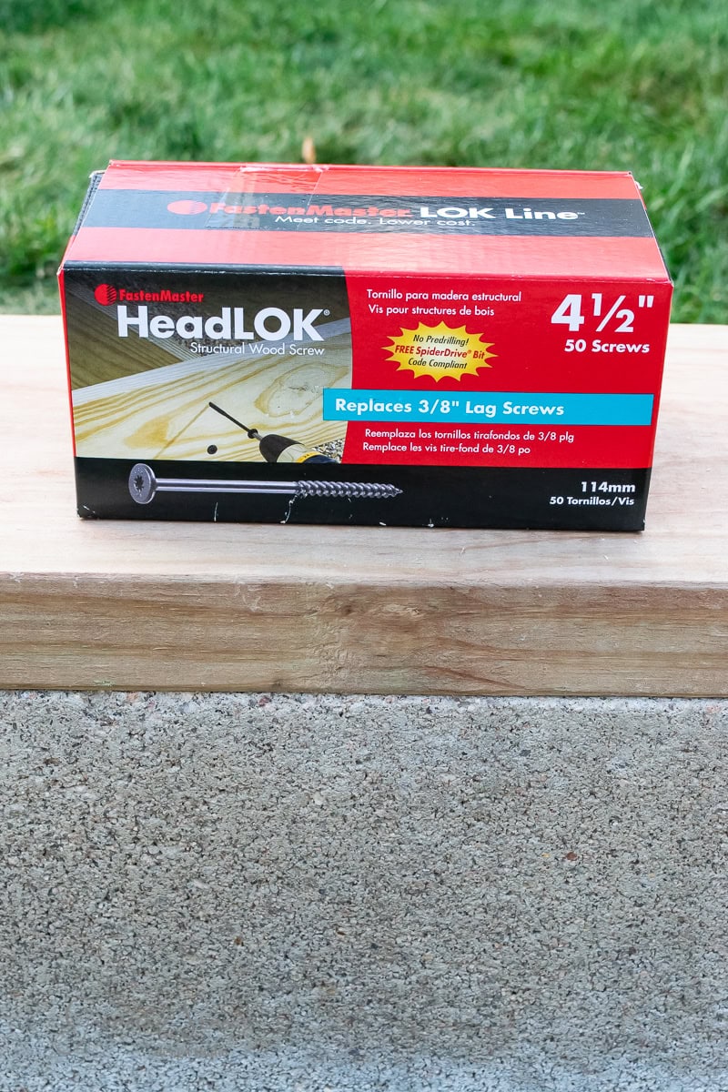 A box of FastenMaster HeadLOK structural wood screws sits on a wooden surface outdoors. The box is red and black, labeled "Replaces 3/8'' Lag Screws" and contains screws that are 4 1/2 inches long.