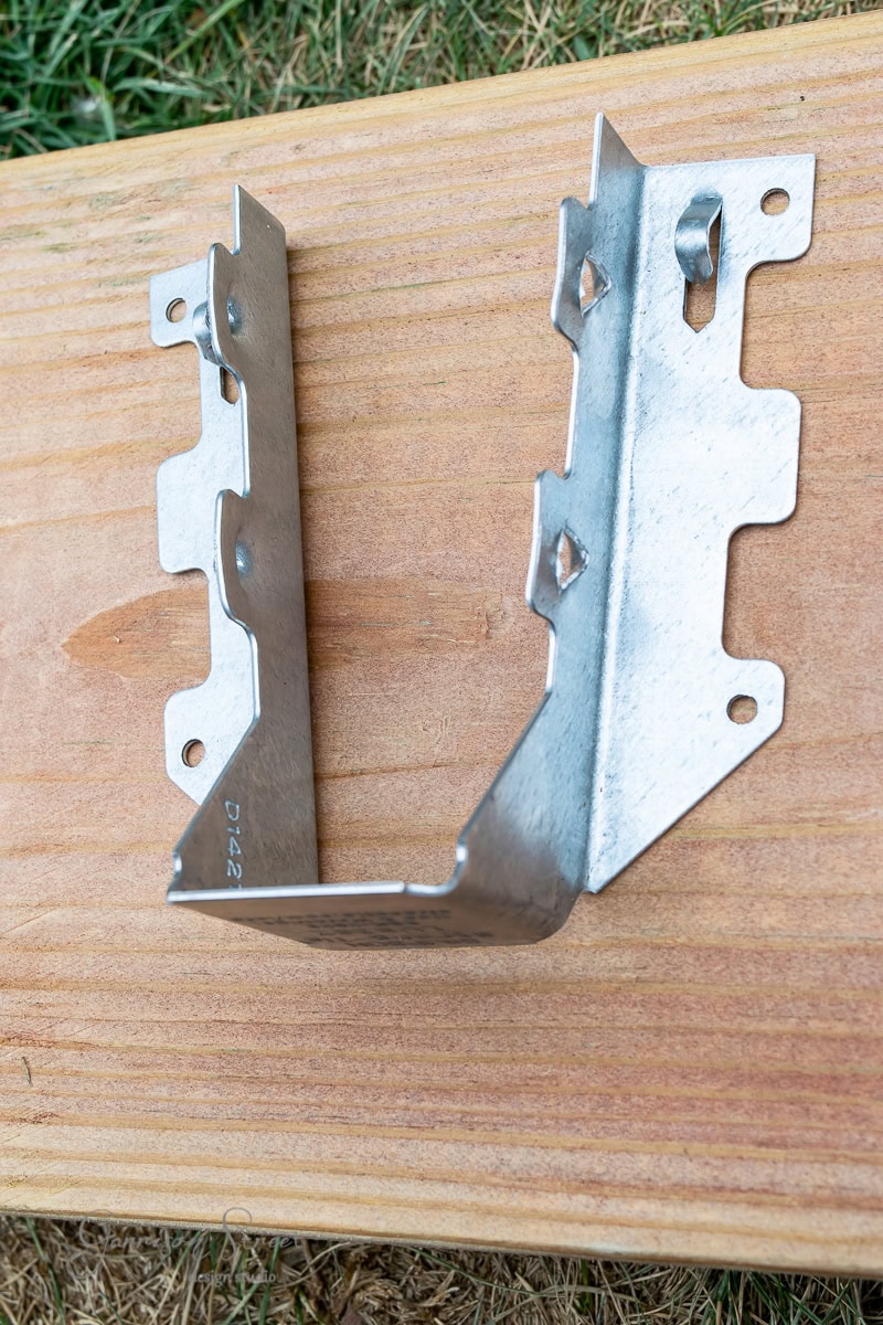 A metal bracket with multiple holes and edges, placed on a wooden surface, likely used for construction or woodworking purposes.