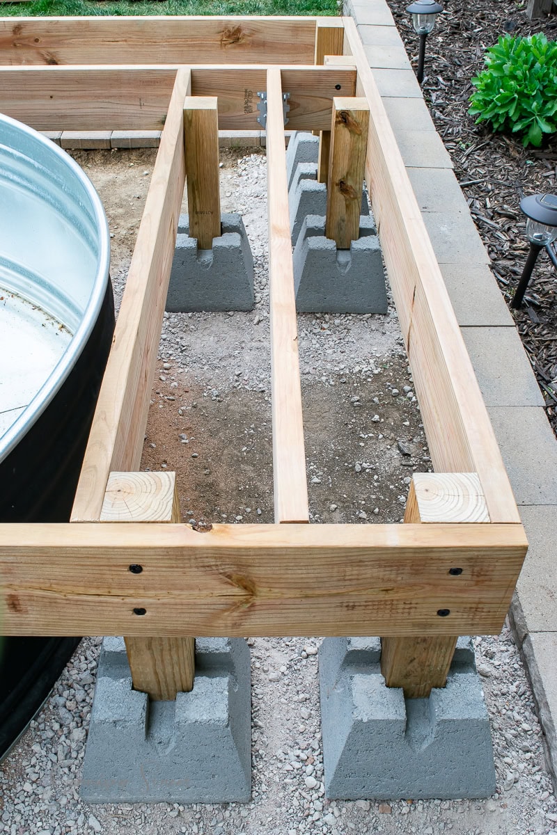 Wooden garden bed frame under construction, supported by concrete blocks. Elevated structure with natural wood beams, set on gravel, surrounded by a garden landscape with green plants and decorative lights.
