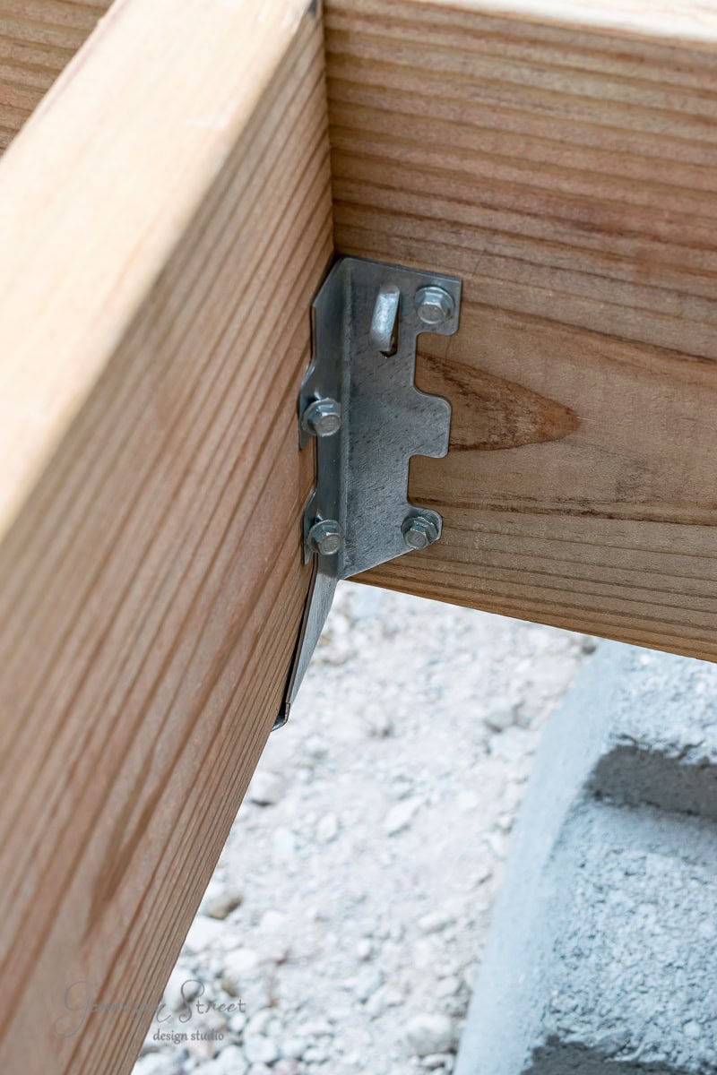 Close-up of wooden beams connected by a metal bracket at a right angle. The beams rest on a textured stone surface. Secure bolts and screws hold the bracket in place, ensuring stability.