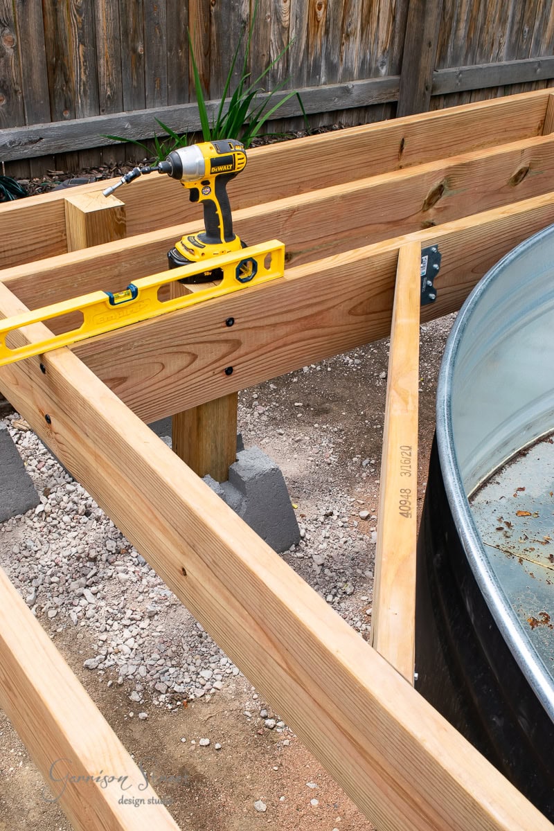 A wooden frame for a deck in progress with a yellow power drill and level placed on the beams. Concrete blocks support the wooden structure. A galvanized metal tub is visible on the right side, and a wooden fence is in the background.