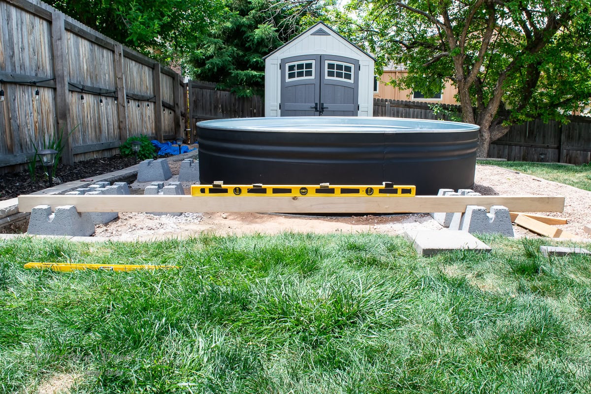 A backyard scene with a round, black above-ground pool on a platform. A yellow spirit level is placed on wooden beams to check alignment. There's a gray shed and trees in the background, with surrounding grass and a wooden fence.