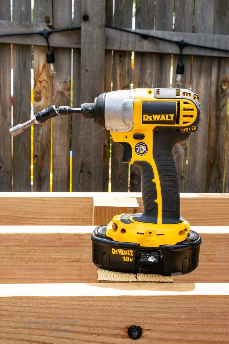 A yellow and black DeWalt 18V cordless impact driver with a drill bit, resting on wooden beams in front of a wooden fence. The tool is in direct sunlight.