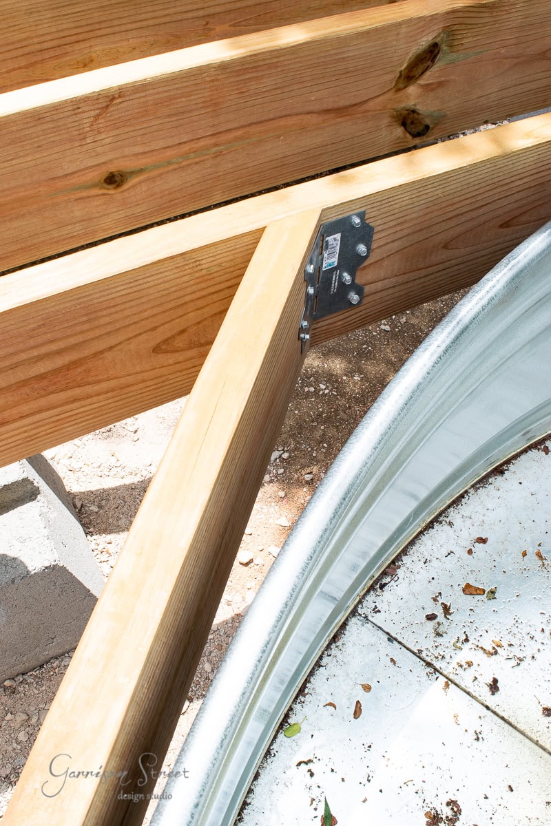 Close-up of wooden beams connected by a metal bracket, part of a construction project. Below is a curved metallic structure on a tiled surface, with some leaves and debris scattered around.