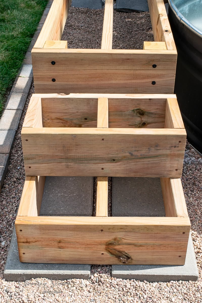 Wooden garden beds are set on a gravel path. The larger bed is behind a smaller, tiered planter. The beds are made of light brown wood with visible knots and joints, placed on gray stone tiles. Grass is visible on the left.