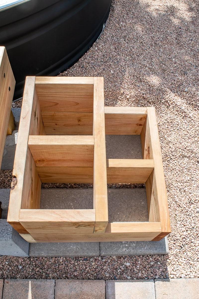 Wooden compost sifters are placed on a gravel surface with sunlight casting shadows. The sifters have wire mesh bottoms and sturdy wooden frames, used for filtering compost.