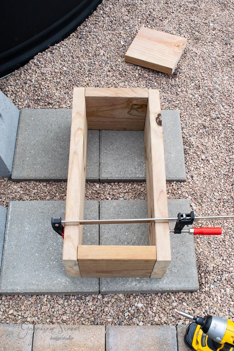 A wooden planter box in the process of being assembled is clamped together, resting on a concrete paver surface. A drill with a bit is placed nearby, and wood planks and gravel are visible in the background.
