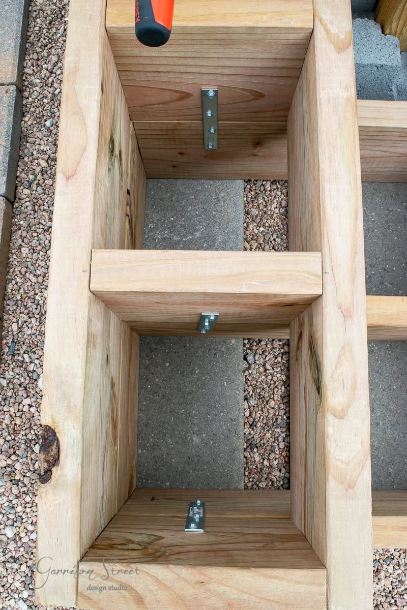 Close-up of a wooden structure being constructed outdoors, featuring interlocking beams and metal brackets for support. The structure is surrounded by gravel, with a few tools visible on the work surface.