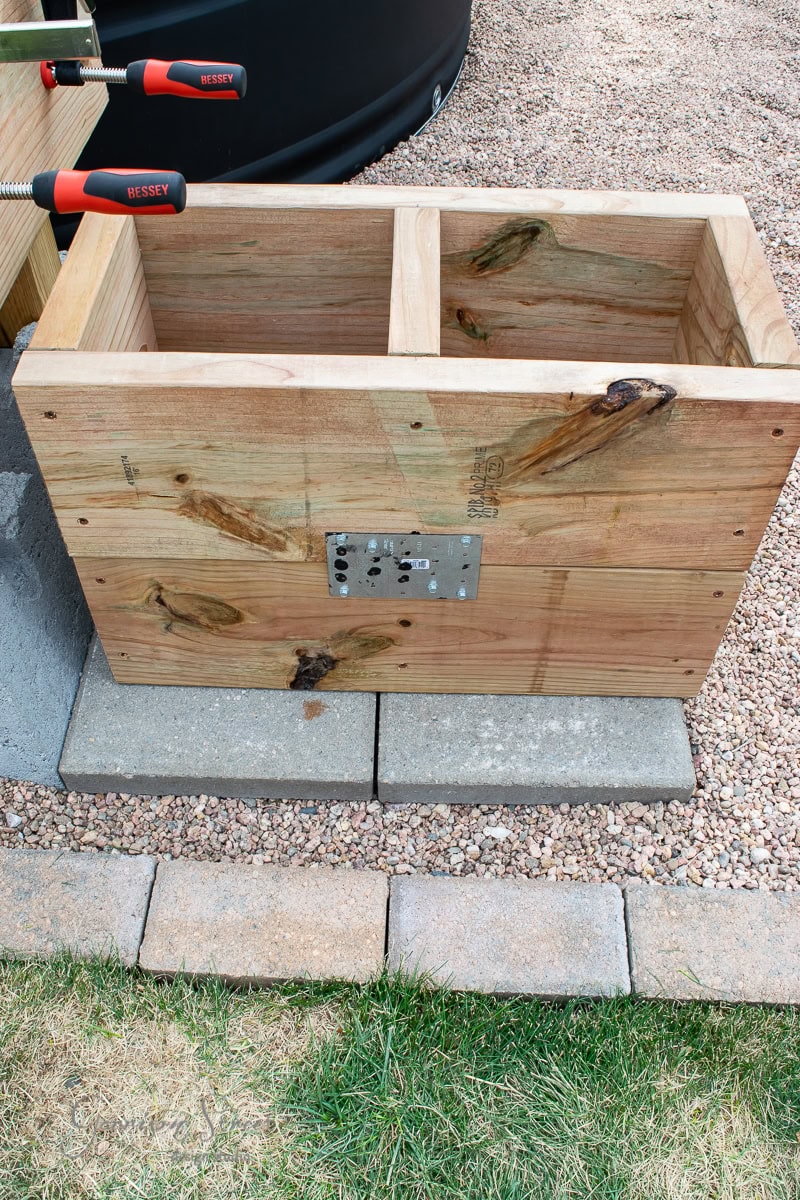 A wooden rectangular planter box, unfinished, with two compartments, sits on a paved surface in a garden. Clamps and carpentry tools are on the edges, and the planter has visible knots and a metal bracket attached to the side.