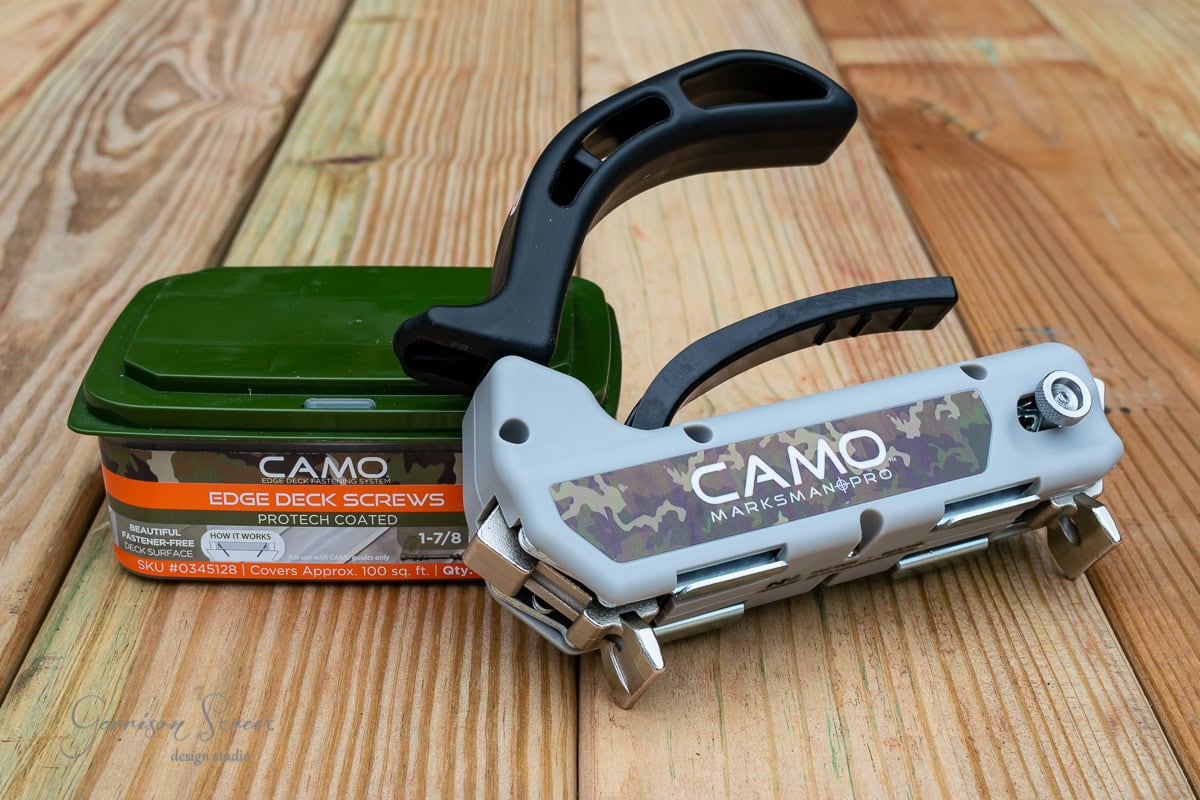 A CAMO Marksman Pro tool lies on a wooden deck next to a green box of CAMO edge deck screws. The tool features a camo pattern and black handle, designed for precise screw placement for decking projects.