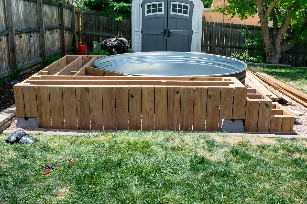 A backyard scene with a galvanized metal stock tank pool placed within a wooden deck structure. The deck has a small set of steps on the right. Tools and construction materials are visible on the grass, and a gray storage shed is in the background.