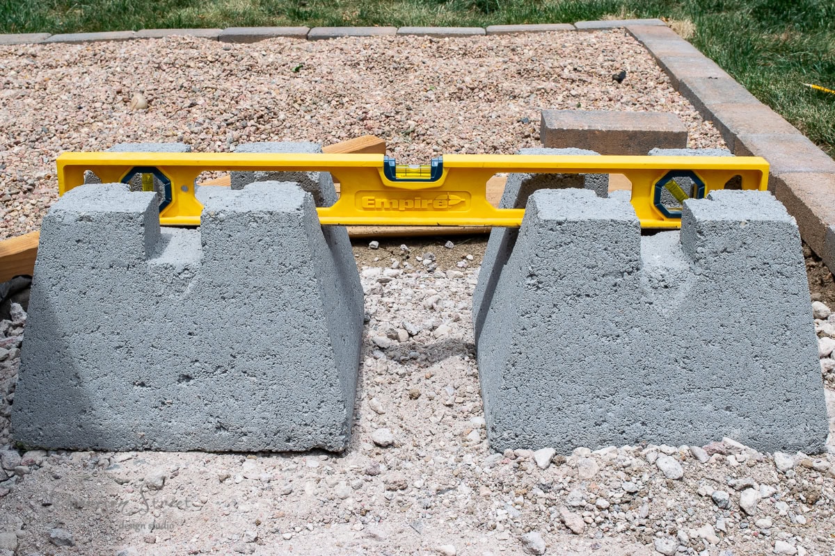 A yellow level is placed across two concrete blocks on a gravel surface, ensuring they are even. In the background, there is more gravel and a partially visible brick border.