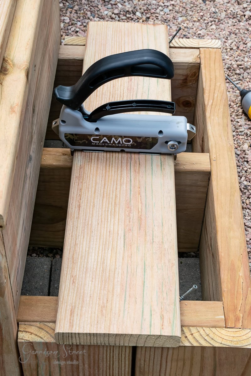 A handheld deck board fastening tool is placed on a wooden plank secured within a rectangular wooden frame. The scene is set on a gravel surface, showing construction or assembly of wooden decking.
