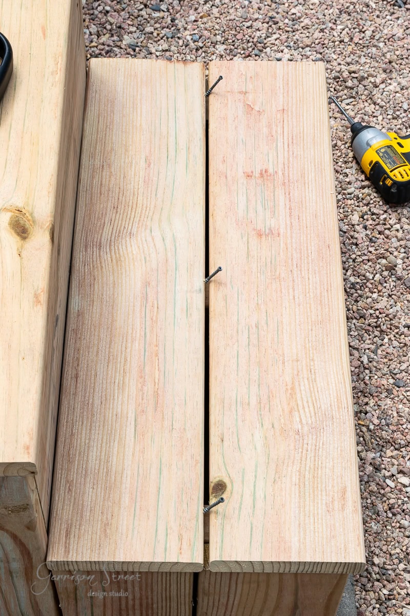 A wooden structure is being assembled with two planks and screws on a gravel surface. A yellow cordless drill lies nearby on the ground.