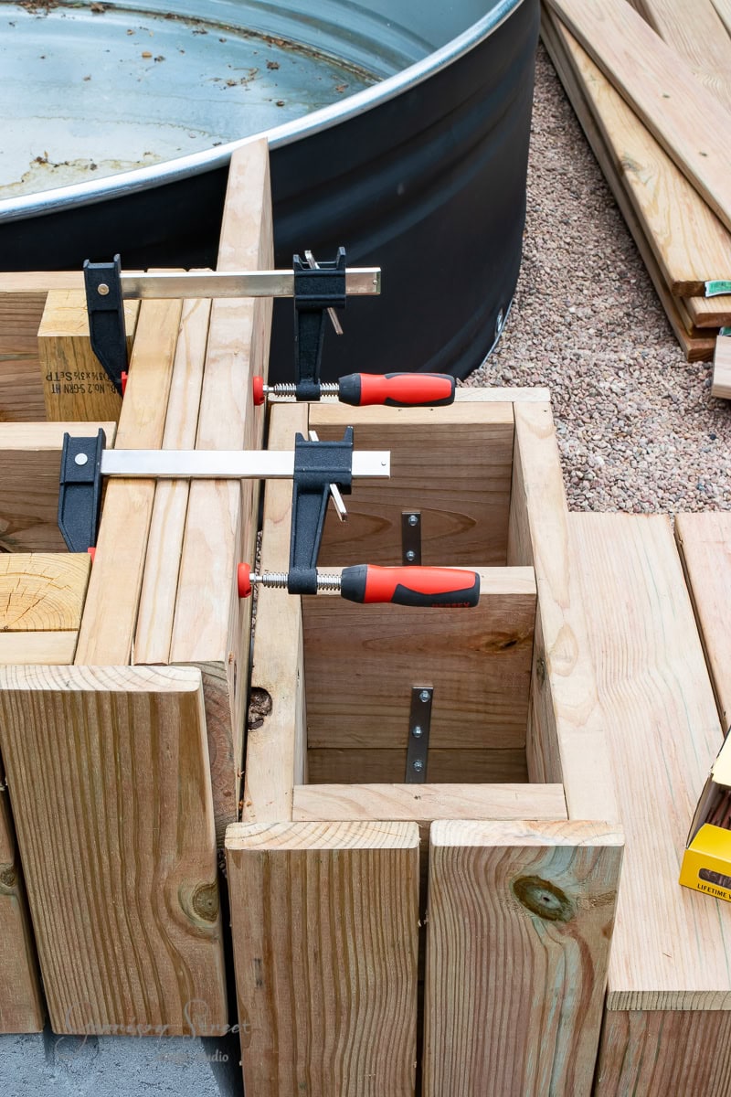 Wooden boards are clamped together, forming a rectangular structure. Metal braces and screws hold it in place. Behind it, there is a large circular metal container and a pile of wooden planks on gravel.