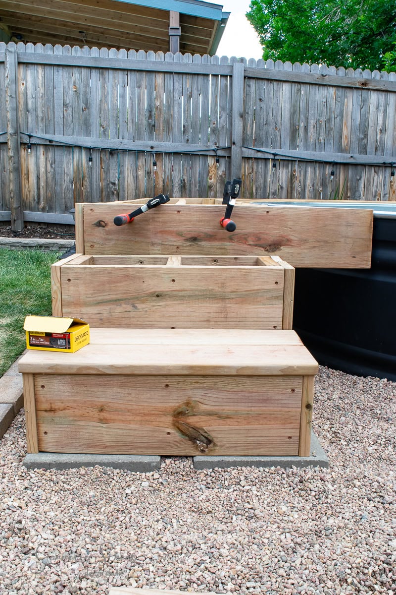 Three wooden planter boxes in a backyard under construction. Clamps are attached to the top box. A box of screws rests on the lower box. The ground is covered with gravel, and a wooden fence is in the background.