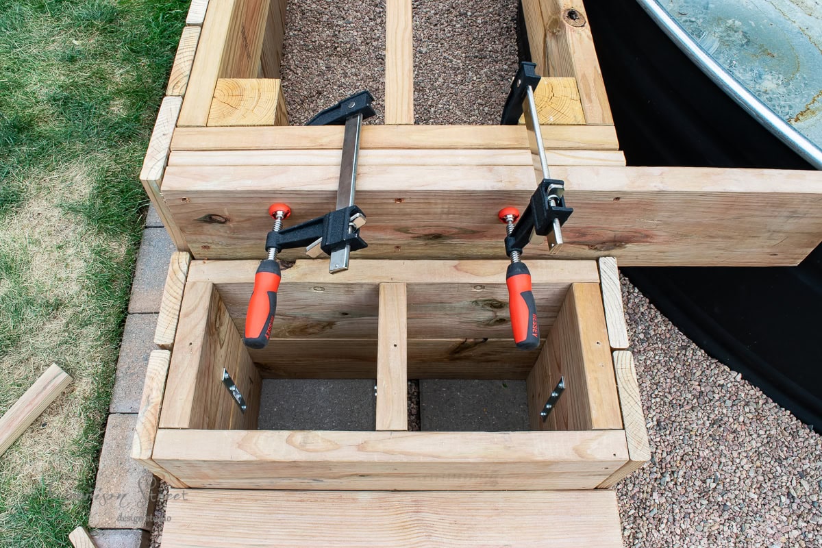 A wooden structure is being built, with clamps holding pieces in place. It is positioned on a lawn next to a gravel area. The construction appears to involve interlocking wood pieces forming compartments.
