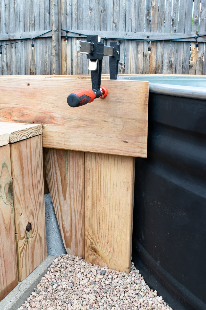 Wooden planks clamped together with a black and red clamp, next to a black metal structure. A wooden fence is in the background, and small gravel stones cover the ground.