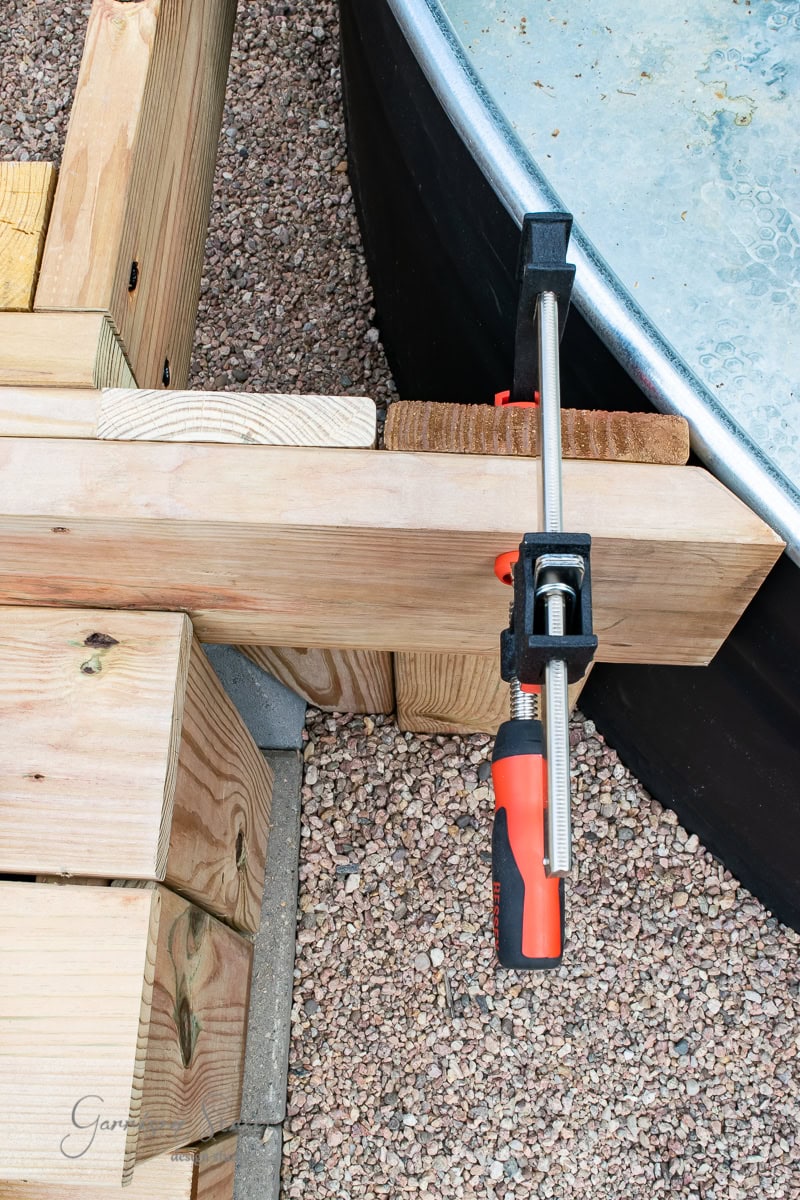 A woodworking clamp secures two wooden beams together on a construction project. The beams rest on gravel, and a large black container is visible in the background. The metal clamp has a red and black handle.