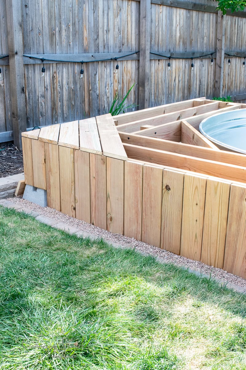 Wooden deck structure under construction in a backyard, adjacent to a lawn and surrounded by a wooden fence. The deck is in progress, showcasing exposed beams and an unfinished surface.