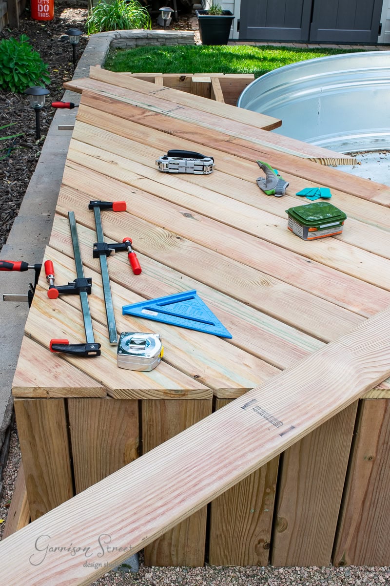 A wooden pool deck is under construction, with a collection of tools like clamps, a speed square, tape measure, and pliers scattered on the surface. In the background, a stock tank pool is partially visible.