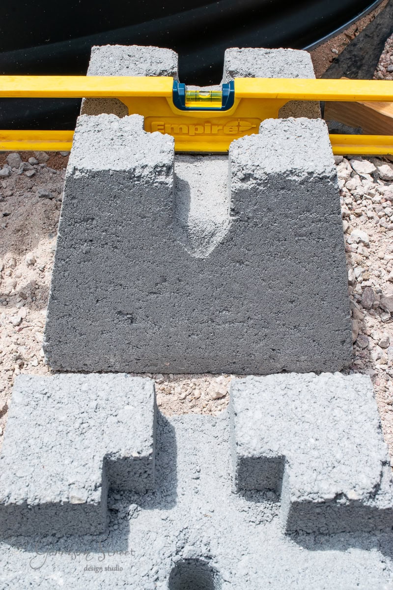 Close-up of concrete blocks with a bright yellow spirit level on top, indicating even alignment. The blocks are on a gravel surface, and the level's bubble is centered, showing precision in construction.