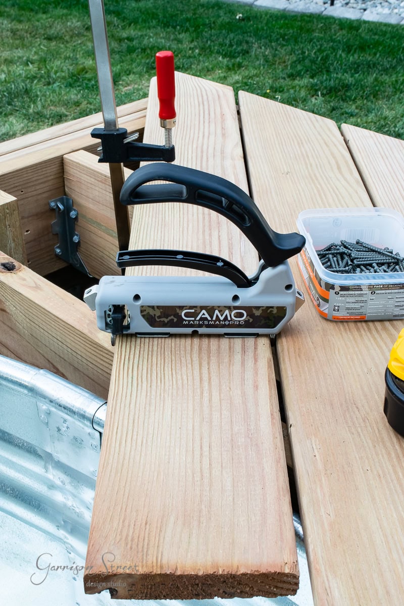 A CAMO deck fastening tool is clamped onto a wooden board on a deck in progress. Nearby are containers with screws and a yellow tool, all set on unfinished decking with grass in the background.