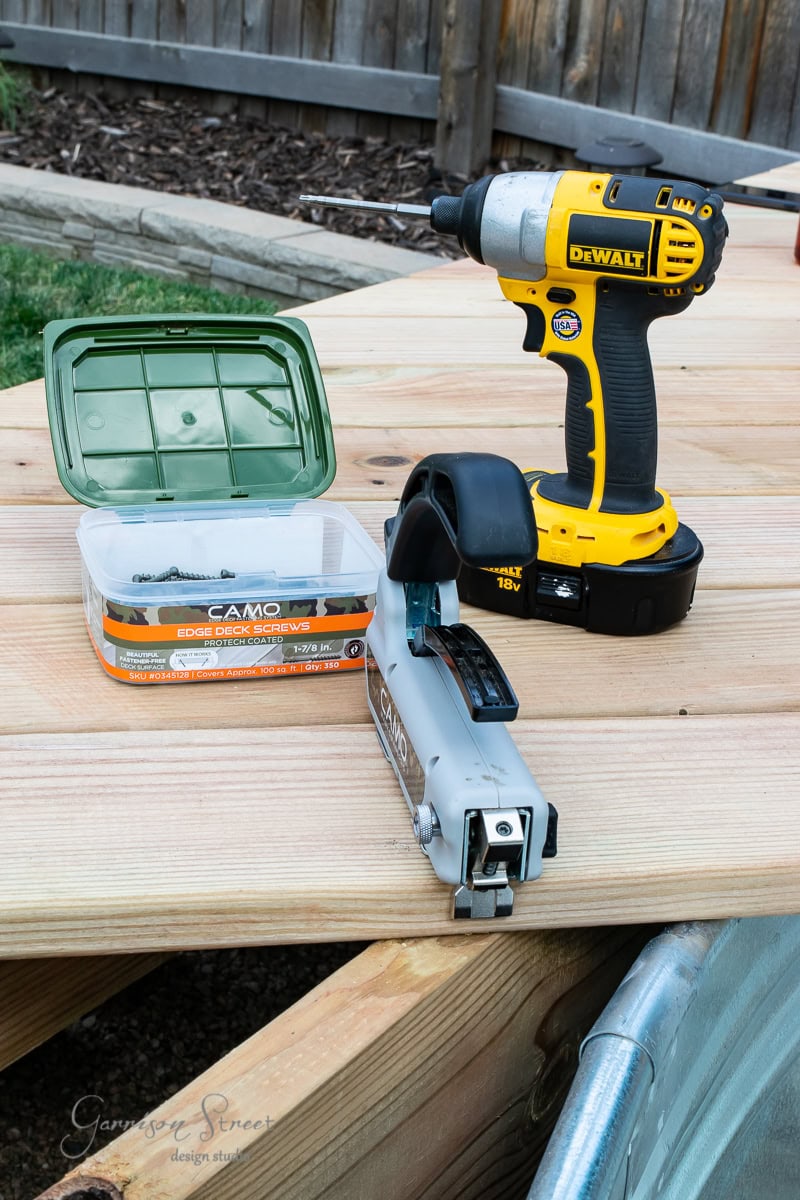 A yellow DeWalt power drill and a green-lidded case of CAMO screws sit on a wooden deck. A CAMO edge deck screw tool is placed nearby. The scene is set outdoors with a wooden fence in the background.