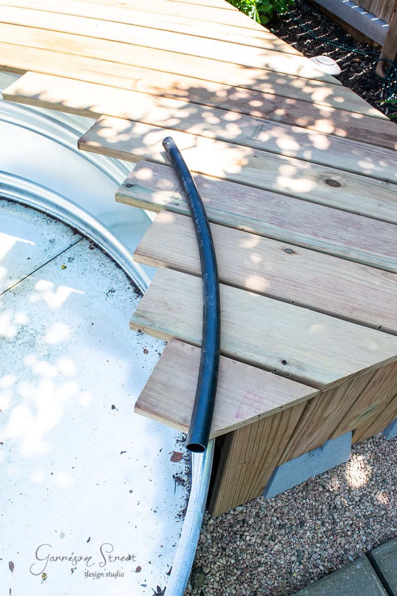 Wooden bench seating curves around a circular metal structure, with a black hose draped across the seating. Sunlight casts shadows on the deck and adjacent garden area, creating a relaxed outdoor setting.
