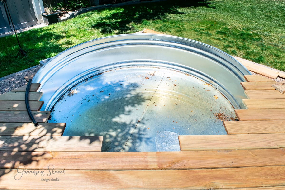 A round stock tank partially dug into a backyard surrounded by wooden decking. The surface of the tank reflects sunlight and has some leaves inside. Grass and shadowed areas are visible in the background.