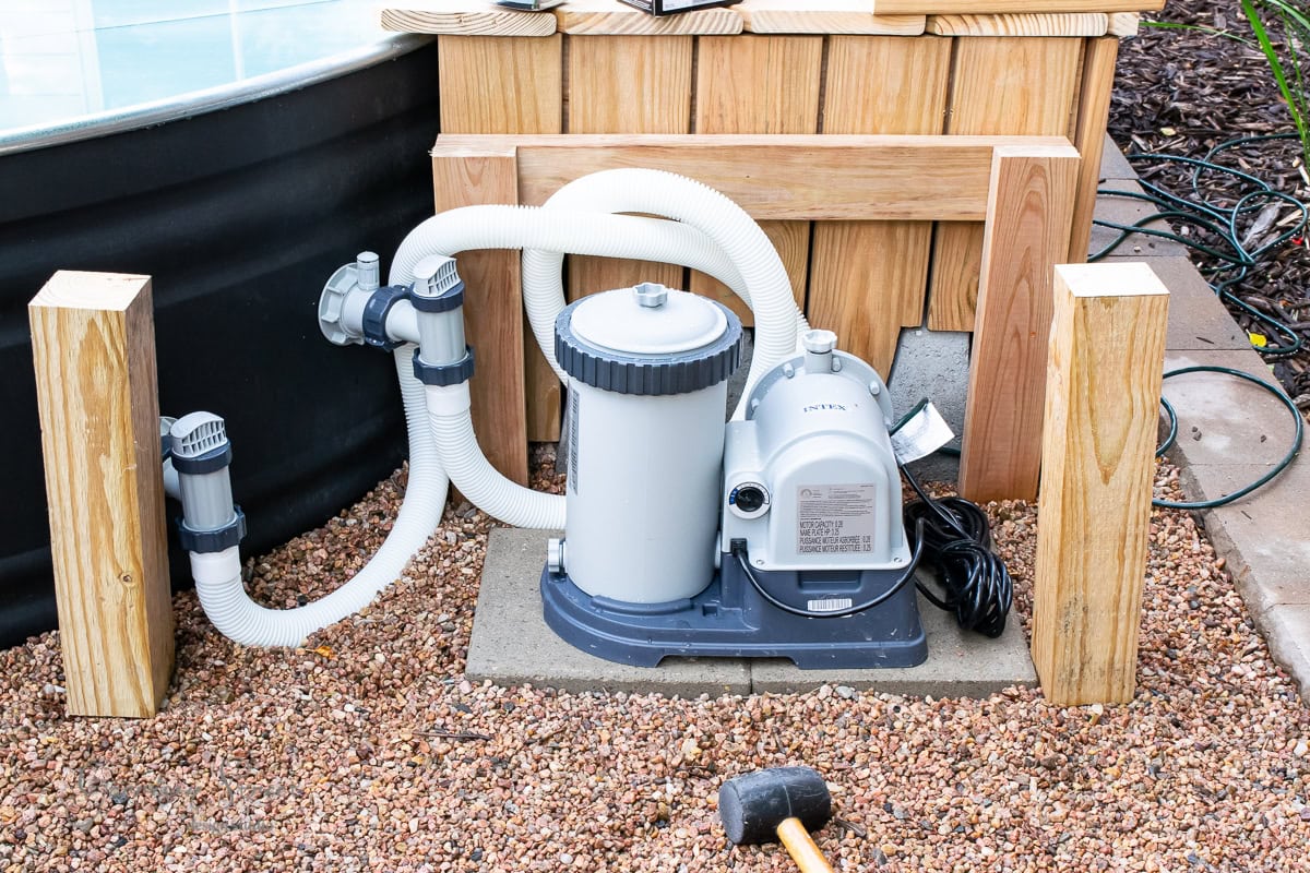 A pool pump and filtration system is set up outdoors on a gravel surface. It is connected by white hoses and flanked by wooden posts. A black rubber mallet lies on the gravel nearby.