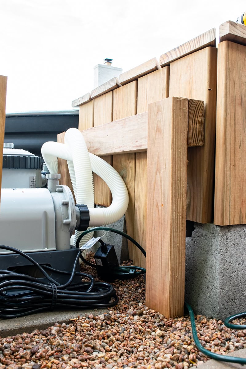 Close-up of a wooden structure on a gravel surface with a white hose connected to a motor or pump. The setup is surrounded by electrical wires. The scene suggests an outdoor plumbing or pool equipment installation.
