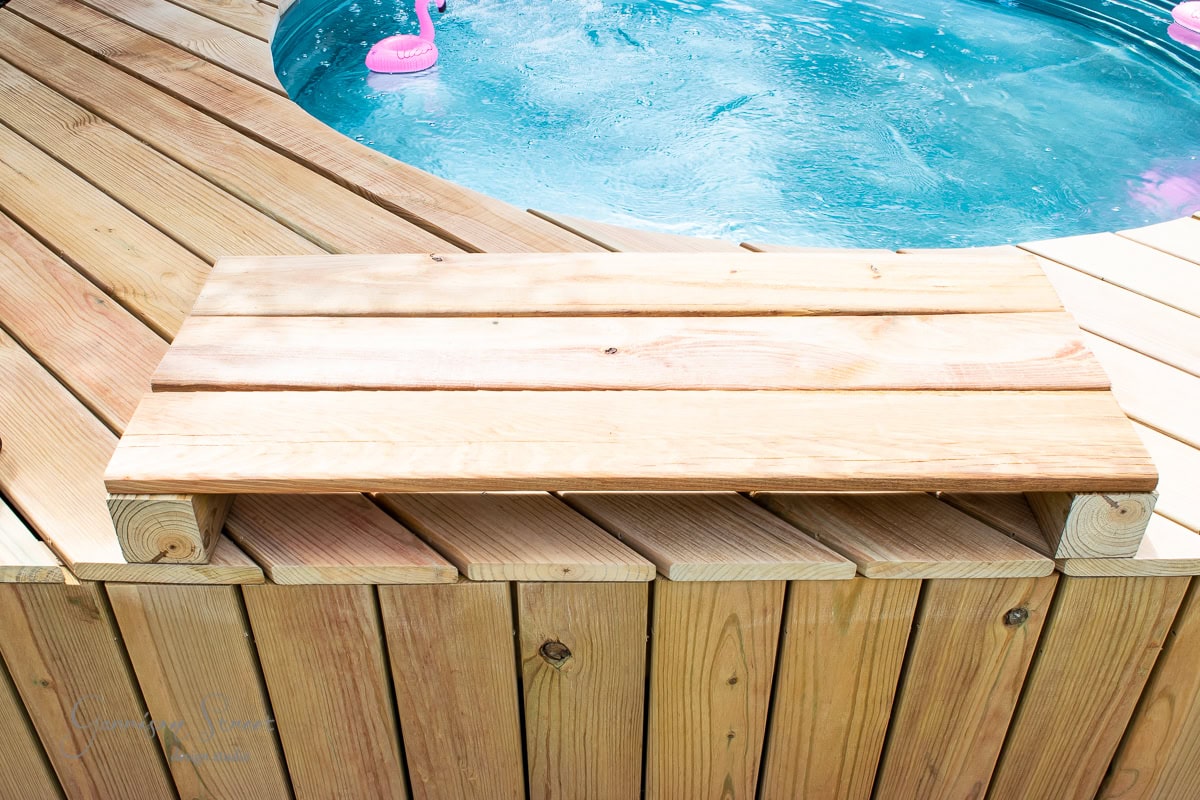 Wooden decking surrounds a circular hot tub filled with bubbling water. Pink flamingo pool floats can be seen in the background. The deck features a simple wooden bench. The scene is bright and inviting, suggesting a relaxing outdoor environment.