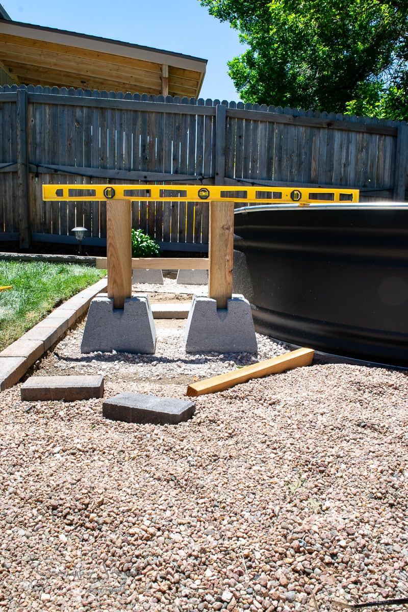 A garden setup with two wooden posts on concrete blocks supporting a yellow leveling tool. They're placed on a gravel area near a large cylindrical container, with a fence and trees in the background. A wooden-handled tool lies on the gravel.