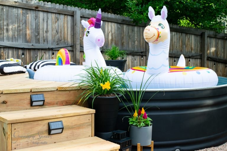 Two inflatable pool floats shaped like a unicorn and a llama are placed in a small backyard pool. The scene is surrounded by wooden decking, potted plants, and a wooden fence in the background. Bright, sunny day.