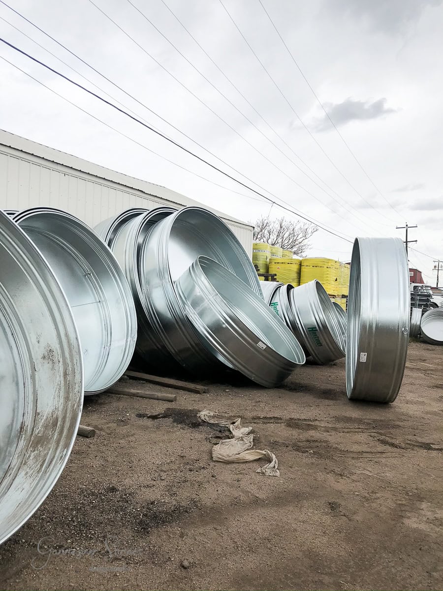 Stacks of large silver metal tanks and lids are arranged on the ground outside. A white building and several yellow boxes are in the background. The sky is overcast with thin, wispy clouds.