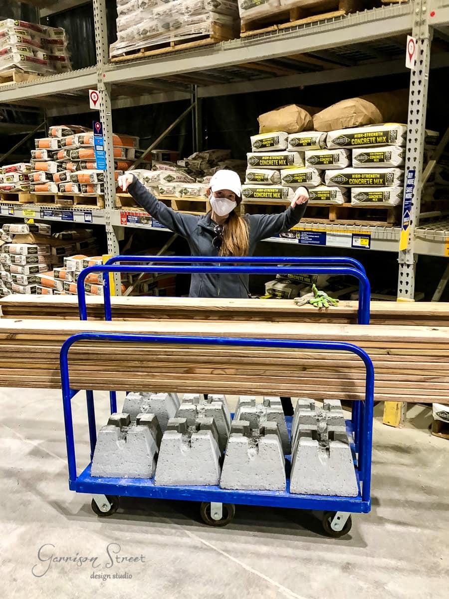 A person wearing a mask and cap poses triumphantly with raised arms behind a blue cart loaded with wooden boards and concrete blocks. Shelves stocked with bags of cement mix and other materials are in the background.
