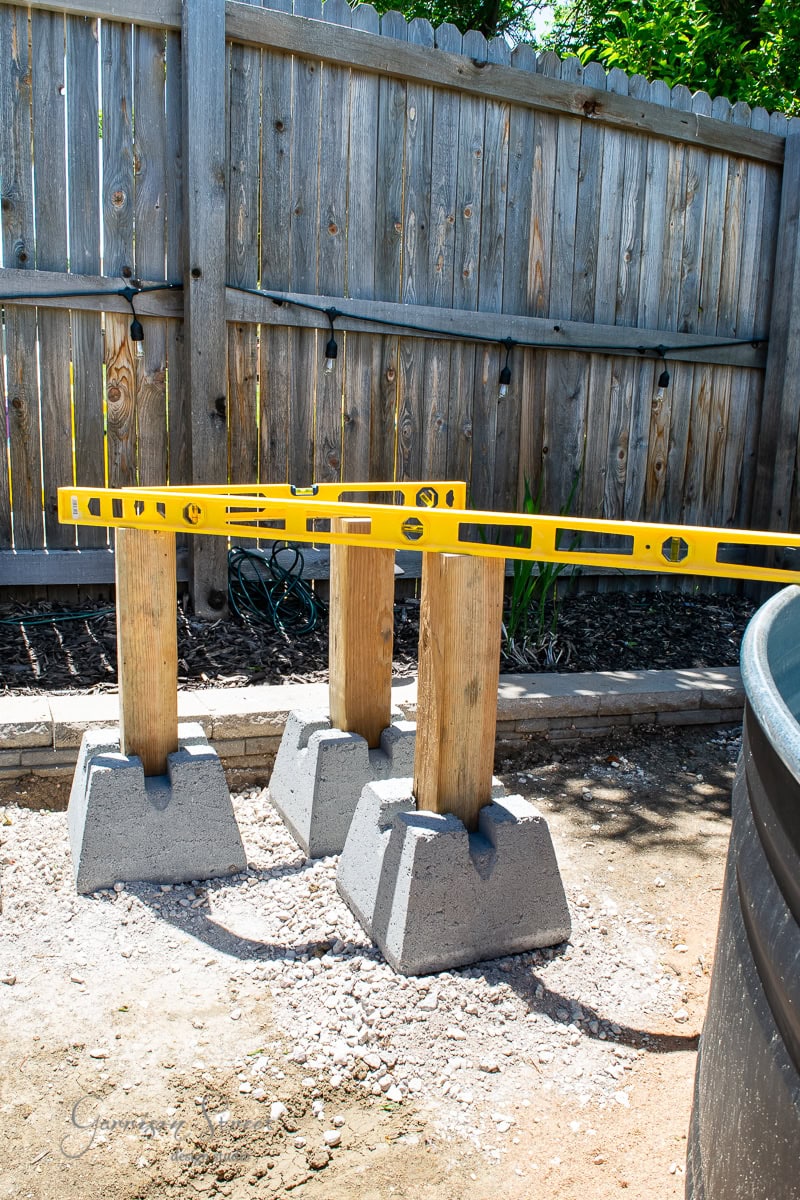 Three wooden posts set in concrete blocks, aligned with a yellow leveling tool on top. The setup is on a dirt surface next to a wooden fence, with plants in the background.