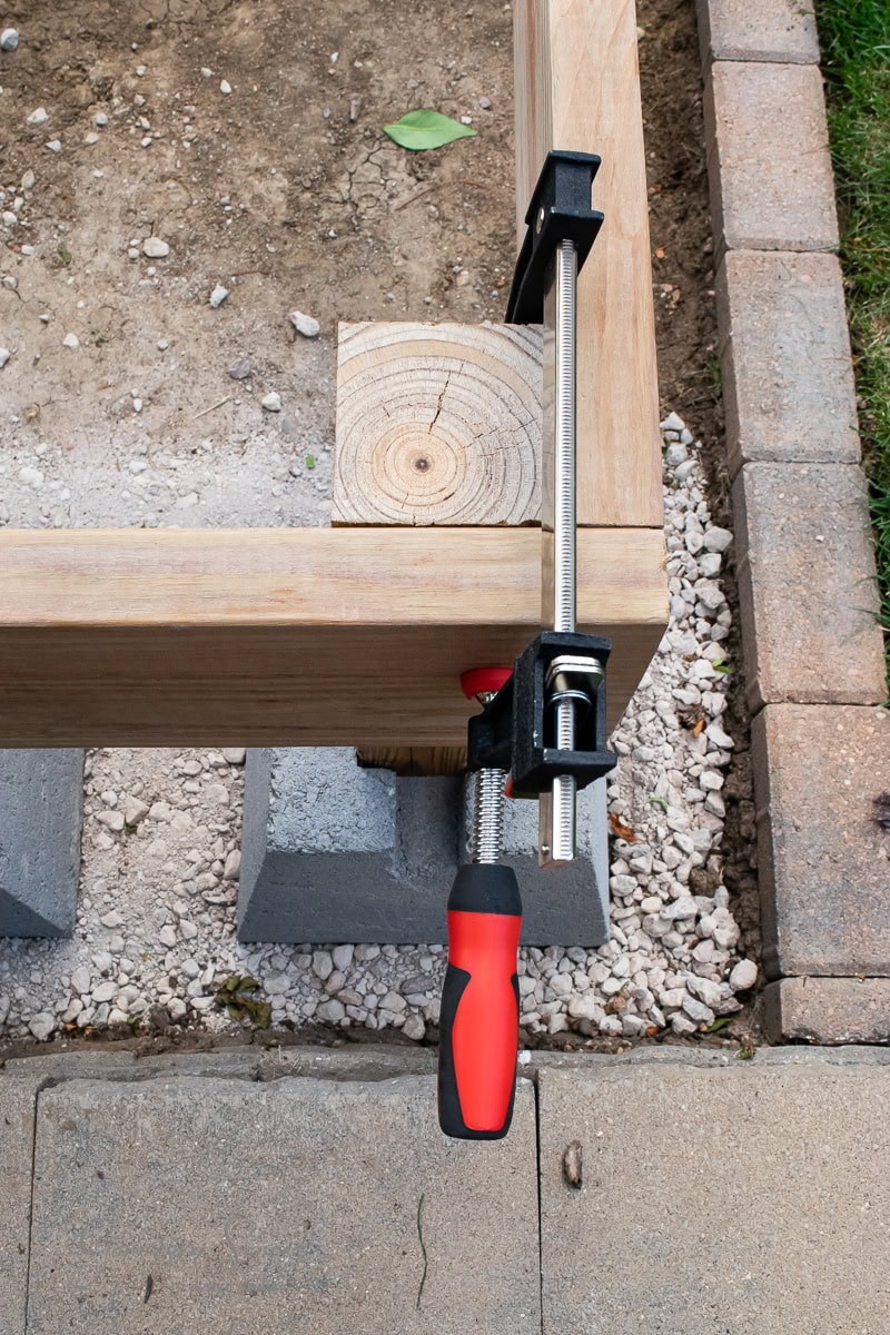 A red and black clamp secures two wooden beams at a right angle. The beams rest on two stacked concrete blocks surrounded by gravel. A stone walkway edge is visible beside grass. A green leaf lies on the gravel.