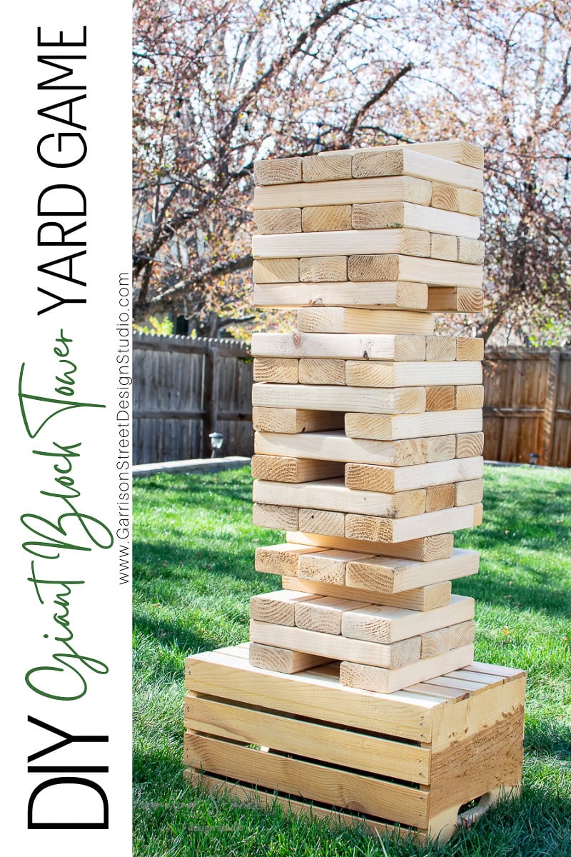 A giant wooden block tower game is set up on a grassy yard, perfect for a summer afternoon. Balanced on a wooden crate, this outdoor game resembles an oversized version of Jenga, with several rows of large wooden blocks. Trees and a wooden fence are visible in the background.