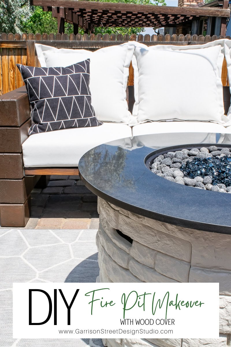 Outdoor seating area with a white cushioned sofa and a black-and-white geometric pillow, perfect for summer evenings. A stone fire pit brimming with blue and gray stones sits in the foreground. The text reads: "DIY Fire Pit Makeover with Wood Cover" and "www.GarrisonStreetDesignStudio.com".