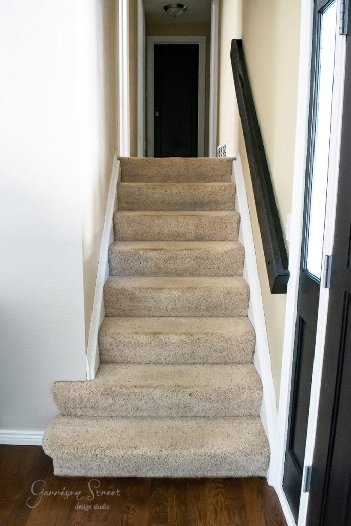 A narrow staircase with beige carpet leads up to a dark door, its stair treads accented by white walls and slightly worn risers. A dark wooden handrail runs along the right, and a wooden floor can be glimpsed at the bottom of the stairs.